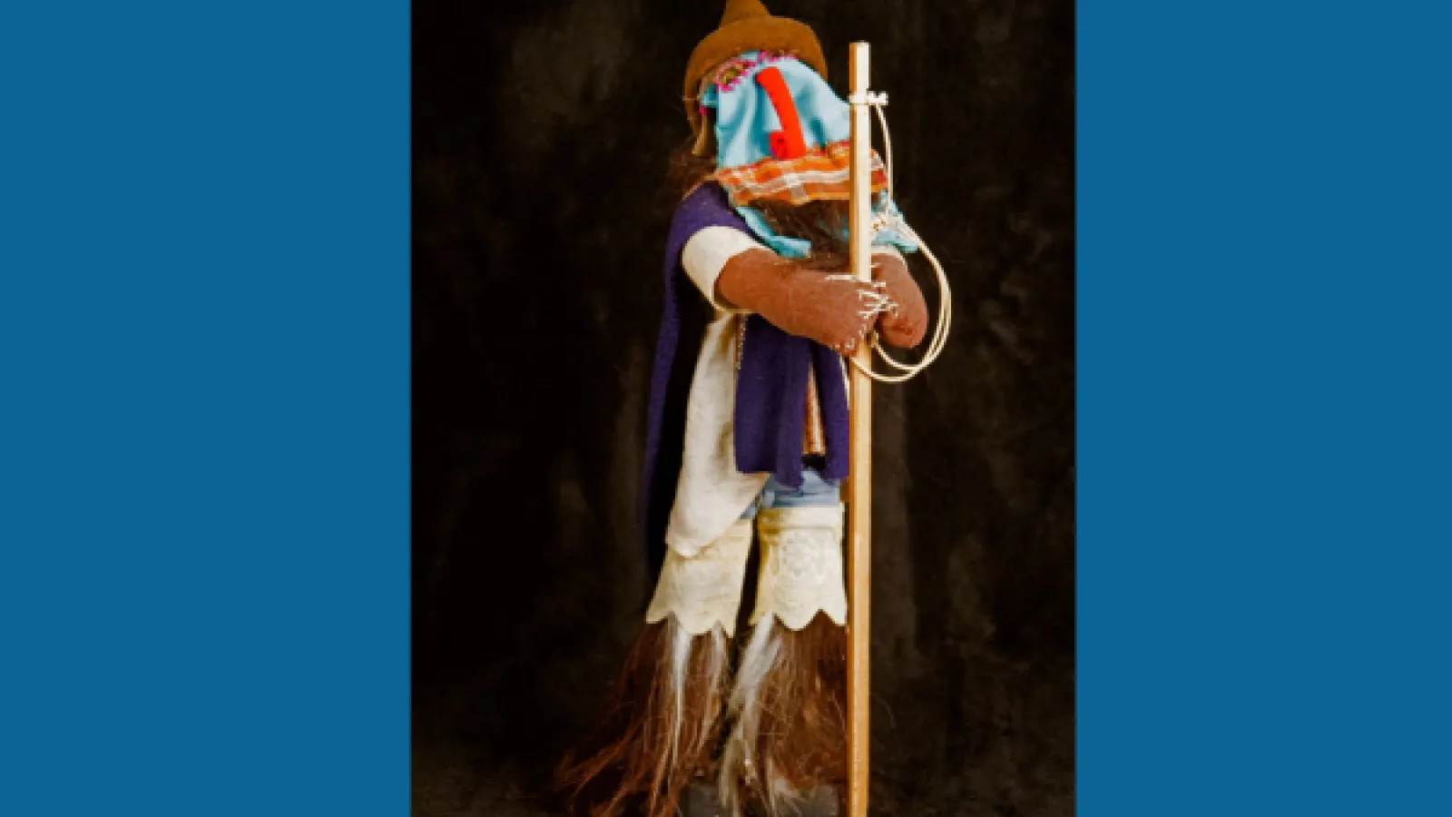 A cloth figure wears a mask and stands on legs made of feathers
