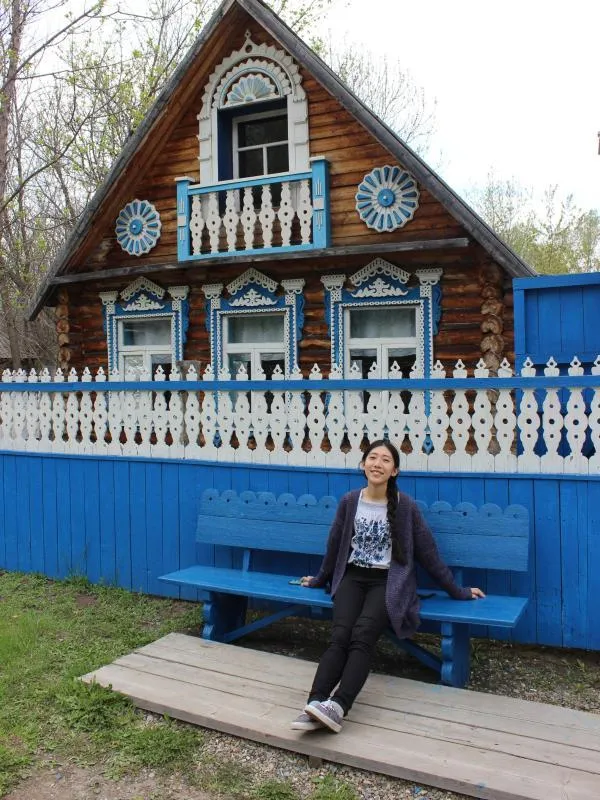 Di Wang sitting on a blue bench in front of a blue and white fence and a wooden house
